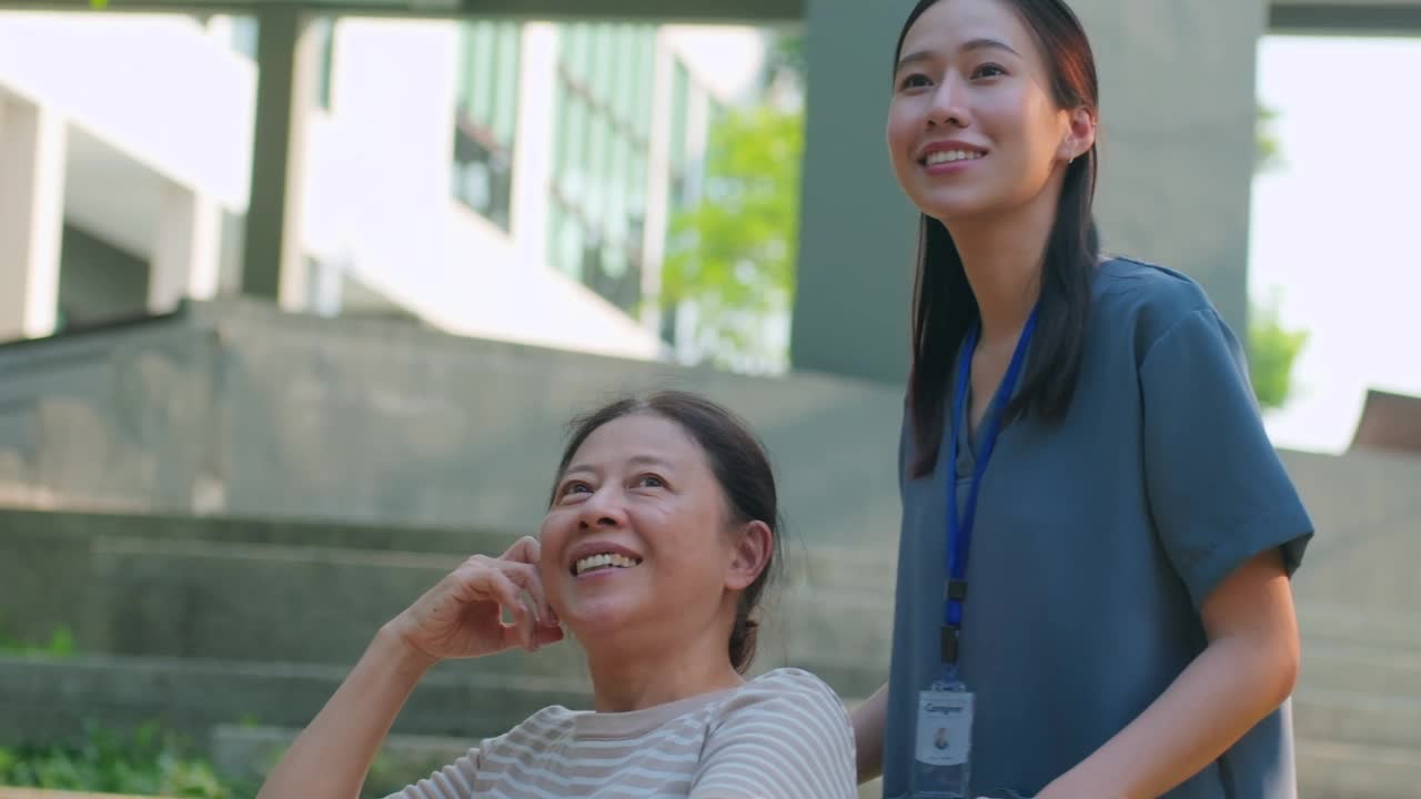 一名身穿蓝色工作服的亚洲护工推着一位坐在轮椅上的老妇人穿过公园，让她呼吸新鲜空气视频下载