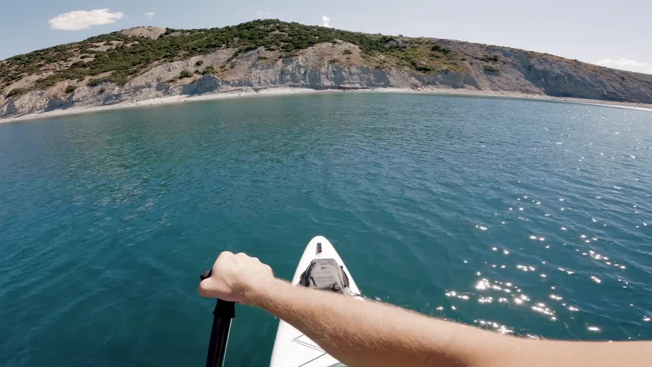 男子在蓝色的海面上划船。第一人称视角的站立桨板在夏季与岩石海岸线。视频素材