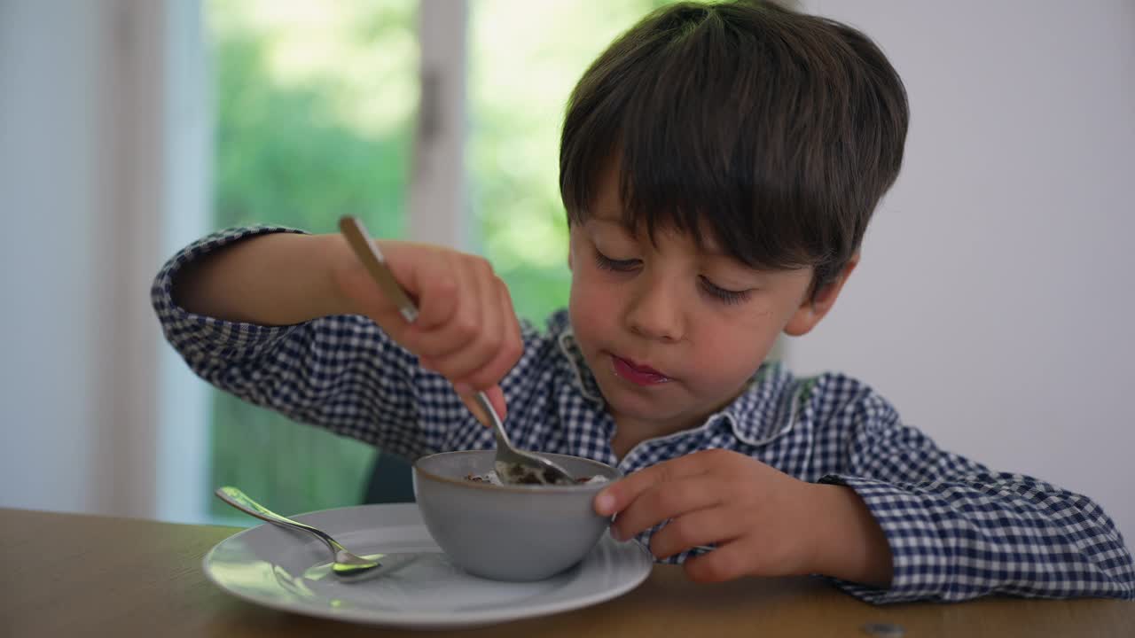 小男孩拿着勺子享用早餐，他坐在厨房的木桌旁，窗外是自然的绿色背景，他表现出专注和享受视频素材