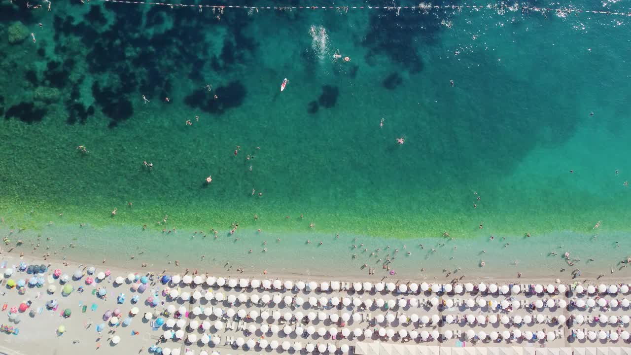 在炎热的夏日，无人机鸟瞰的海滩上挤满了人。假日旺季。白色遮阳伞在完美的沙滩上排成一排。视频下载