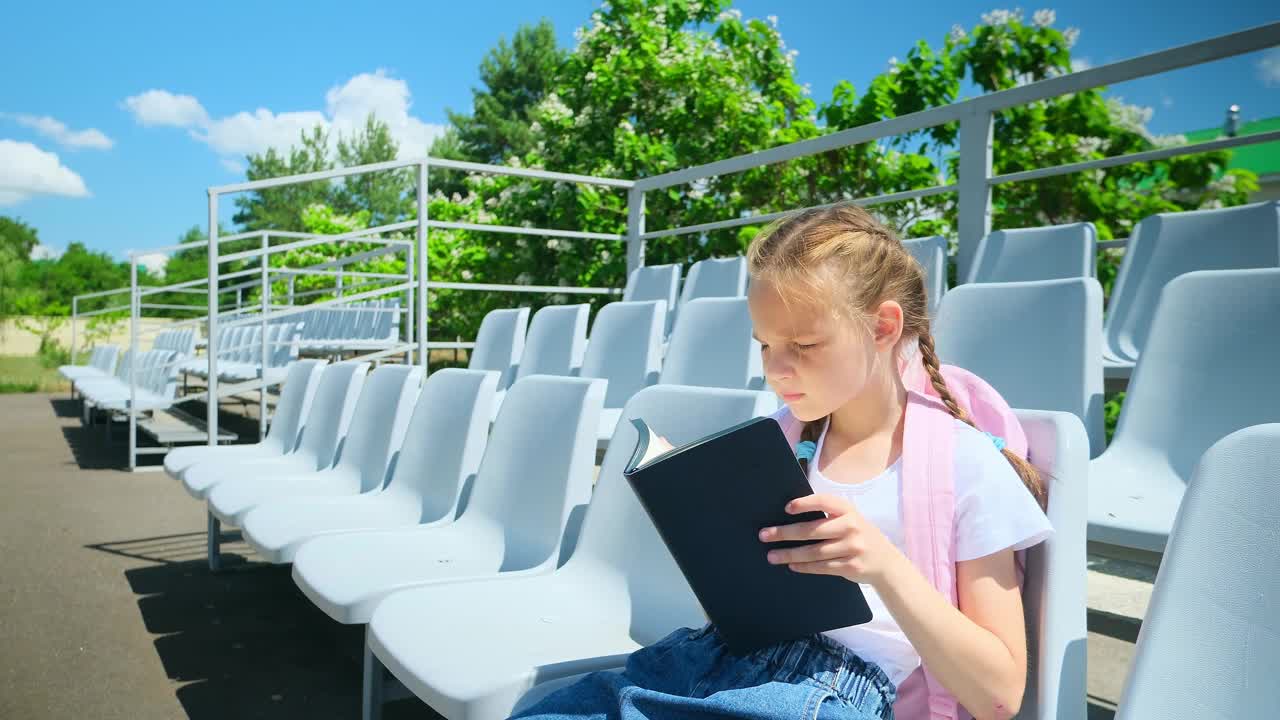 回到学校。小学生。一年级生。开始上课了。一个背着书包的小女孩坐在长凳上，手里拿着课本。视频素材