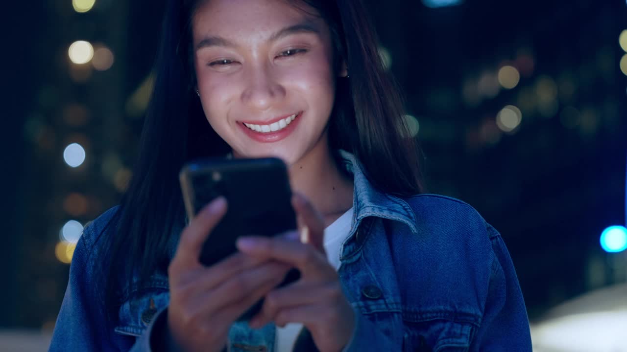 美丽的年轻亚洲女子站在城市街道在晚上使用手机。一个快乐的女人站在那里用手机打字视频素材