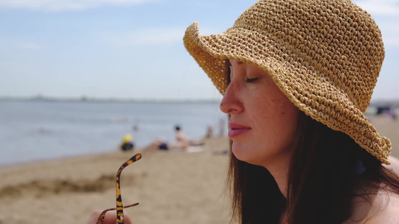 美丽的女旅行者肖像，戴着草帽，戴着太阳眼镜，带着草包环顾四周，走在沙滩上。视频下载