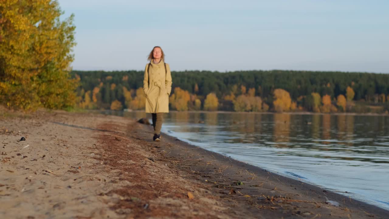 孤独的女人在河边欣赏秋天美丽的日落。穿大衣的女士沿着海岸散步，望向远方。视频素材