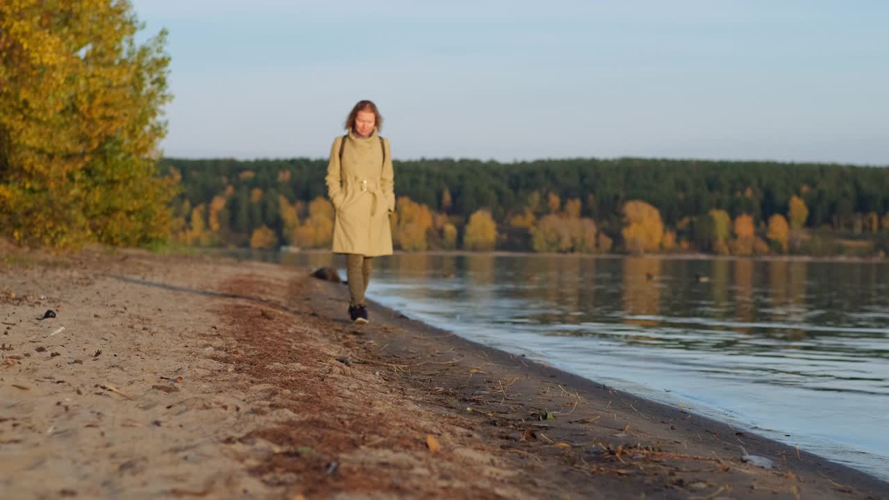 孤独的女人在河边欣赏秋天美丽的日落。穿大衣的女士沿着海岸散步，望向远方。视频下载