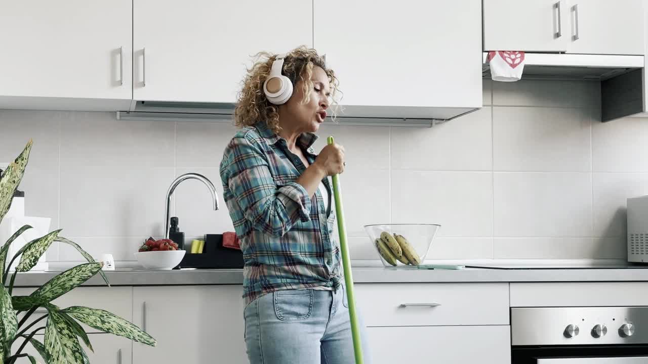 一个兴奋的女人在家里戴着耳机在厨房里跳舞，拿着扫帚唱歌。疯狂自由，心情愉悦的生活方式。卷发的中年女士在公寓里玩得很开心视频下载