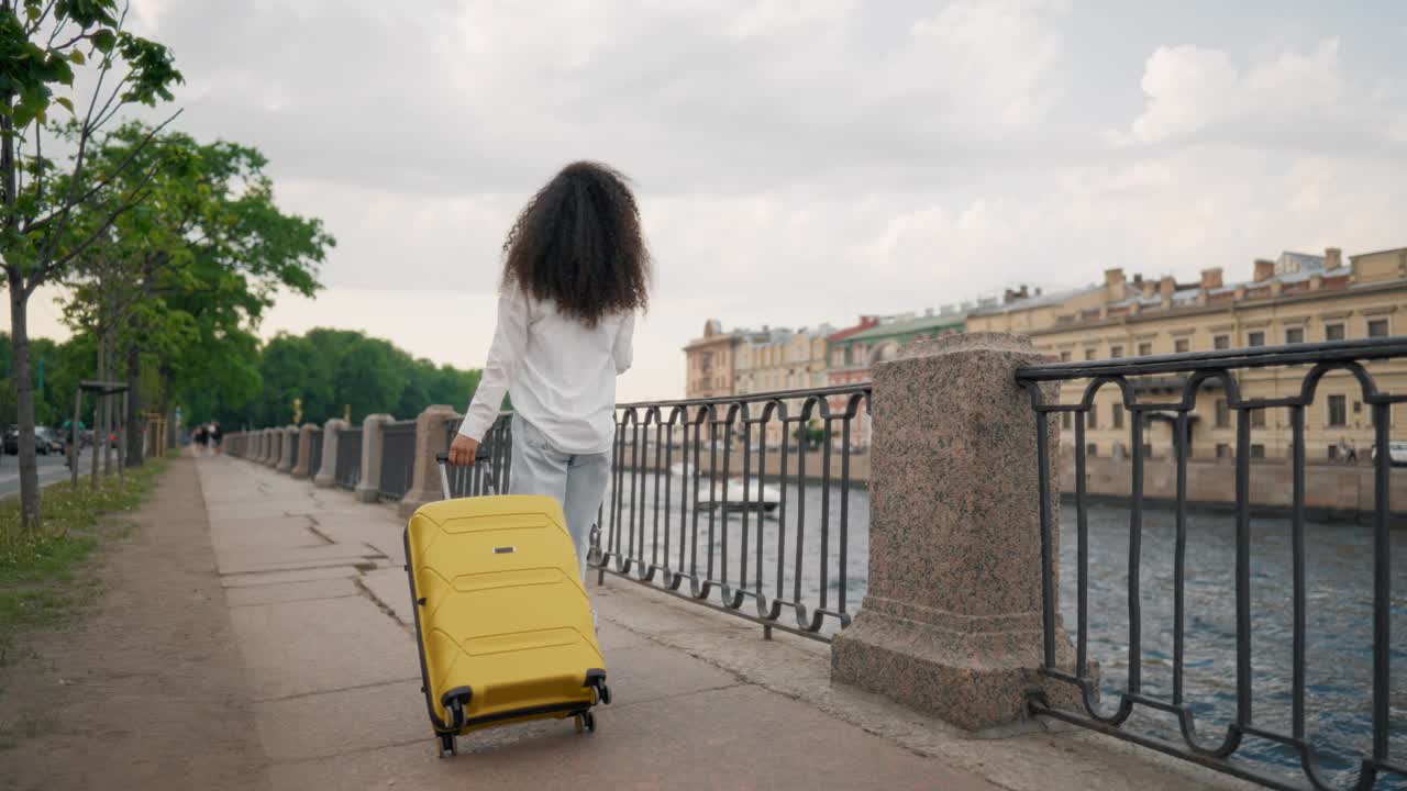 非裔美国人女士带着大黄皮箱参观古老的旅游城市。快乐的西班牙女游客独自走在河堤上的清晨，向后看，观光和旅行到欧洲视频素材