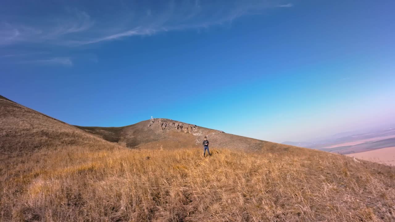 一名戴着FPV眼镜和头盔的白人男子站在山上，控制着一架在自己周围飞行的FPV跨无人机四轴飞行器。无人机驾驶员的动态画面视频素材