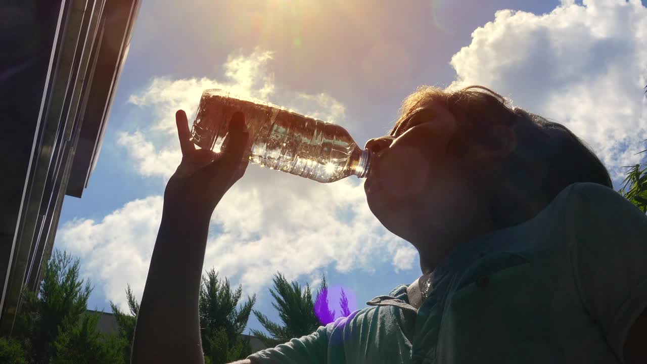 一个男孩在阳光明媚的大热天用瓶子喝水。视频下载