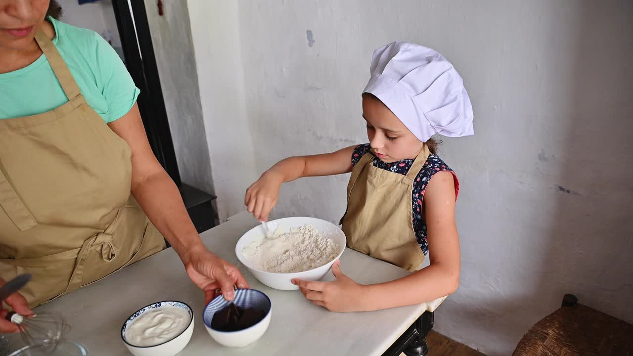 孩子戴着厨师帽，系着围裙，在指导下在厨房里混合食材视频素材