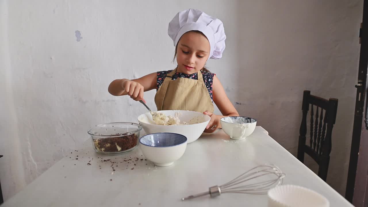 戴着厨师帽系着围裙的小女孩在厨房里搅拌食材视频素材