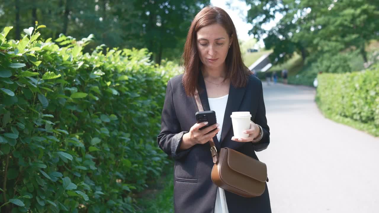 一位女商人正在户外散步，手里拿着一杯咖啡，手里拿着她的智能手机视频下载