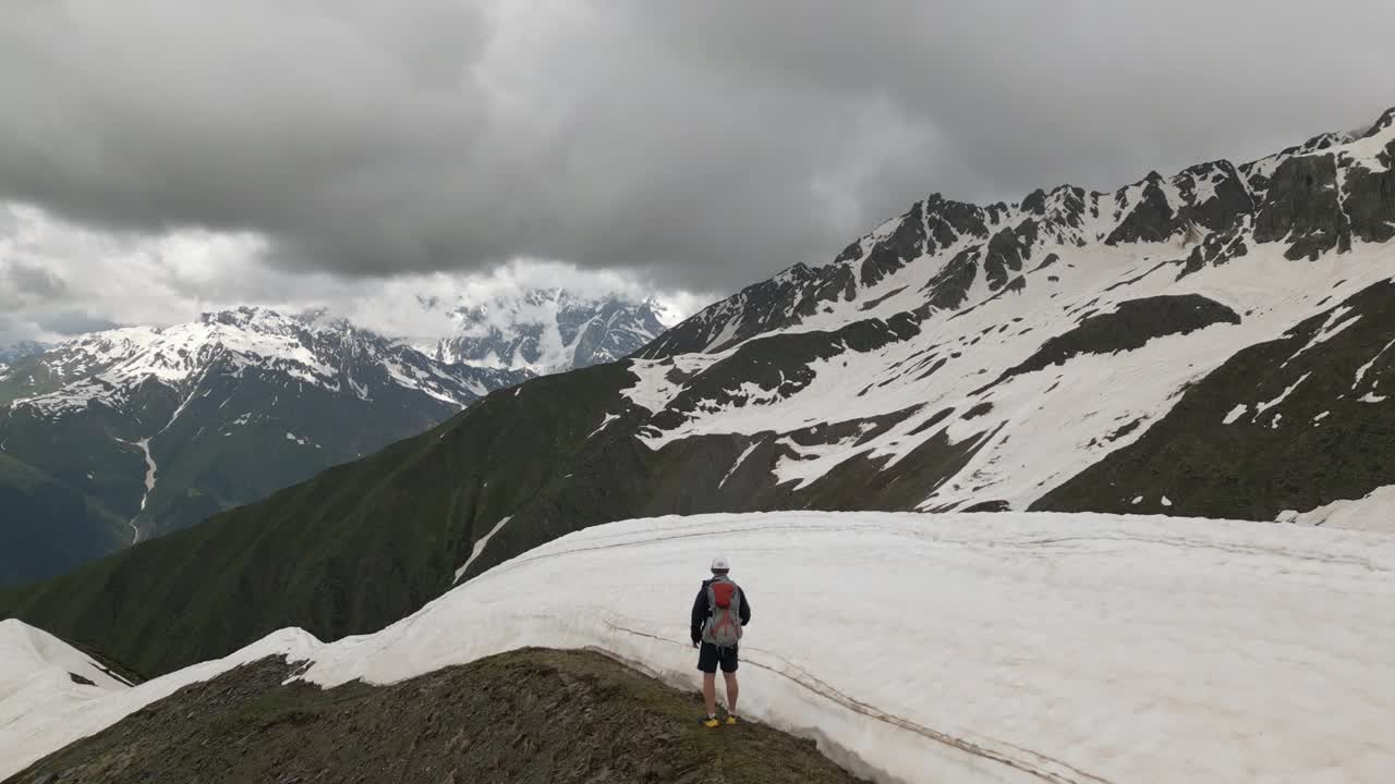 一个独自背包客站在高高的雪山上。鸟瞰图。徒步者被雪包围，山峰崎岖不平，天空多云。户外探险徒步旅行，探索大自然荒野，雄壮的无人机景观视频素材