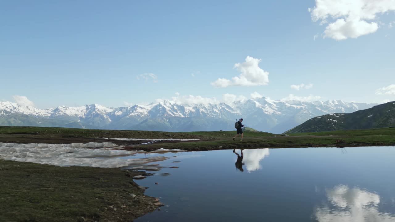 徒步旅行者在斯瓦涅季山脉Koruldi湖边散步的鸟瞰图。一个人独自在大自然中背包旅行。宁静的水面，白雪皑皑的山峰，孤独，自我反省，冒险。无人机拍摄的视频素材