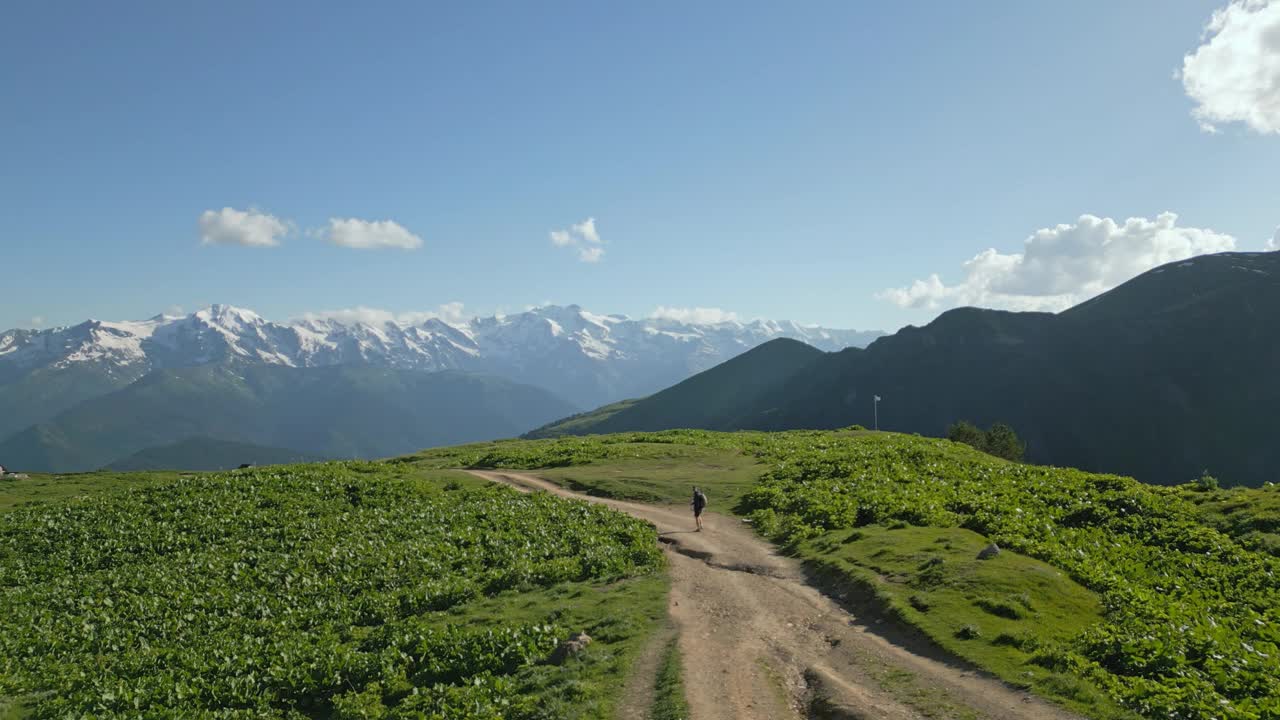 单人背包客徒步穿越高山草甸的航拍图。背着背包的人沿着土路走。青山绿水，雪峰蓝天。无人机拍摄的视频素材