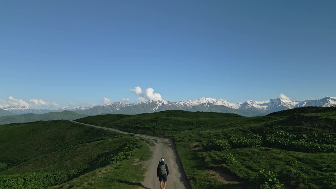 男子在高山草甸小径上徒步。航拍画面显示了广阔的视野，雪峰，晴朗的天空。户外探险、自然探险、徒步旅行。无人机拍摄的视频素材