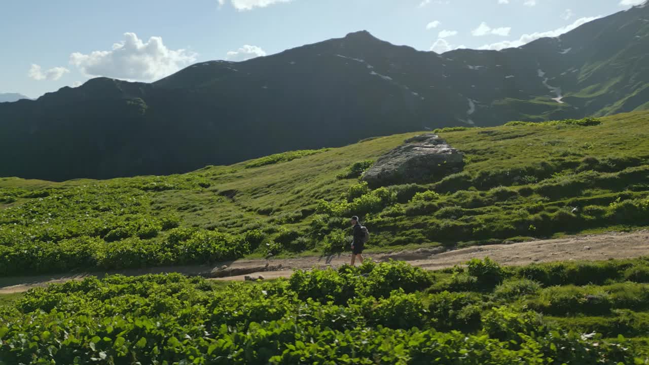 单独的男性旅行者在斯瓦涅季山脉的高山草地上徒步旅行的鸟瞰图。在阳光明媚的夏日，人们探索着绿色的山丘和白雪皑皑的山峰。冒险，自然，徒步旅行。视频素材