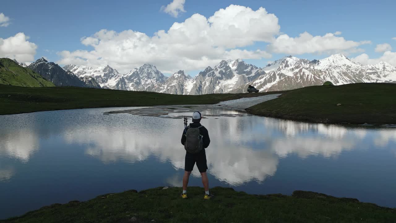 Koruldi湖徒步旅行者背包鸟瞰图。探险家站在水边，映照天空，雪山。山区徒步旅行，冒险，自然探索，斯瓦涅季地区。无人机拍摄的视频素材