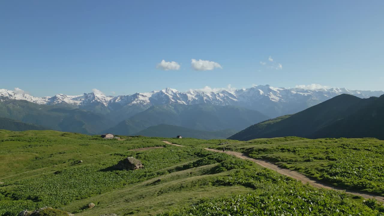 在山脉中徒步穿越高山草甸的航拍照片。徒步旅行者走在绿色田野和雪峰环绕的小路上。大自然的探险冒险，崎岖不平的风景，湛蓝的天空。视频素材