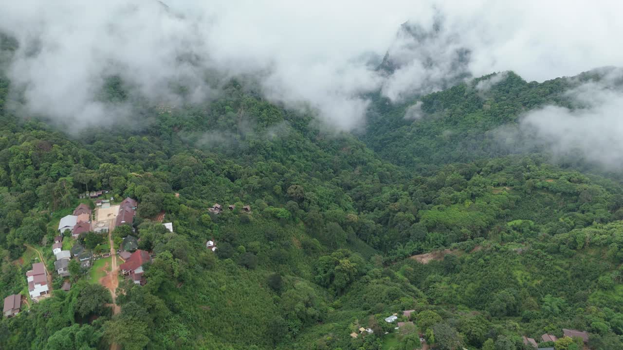 空中拍摄的茂密的绿色森林覆盖了山区地形，山谷中有一个小村庄，被雾和云包围着。视频下载