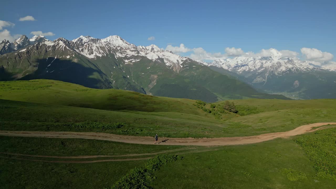 鸟瞰图上的男子徒步旅行的绿色高山草地在高山雪峰。孤独的徒步者走在泥泞的小路上。探索自然和遥远的荒野。天气晴朗，山景优美。视频素材