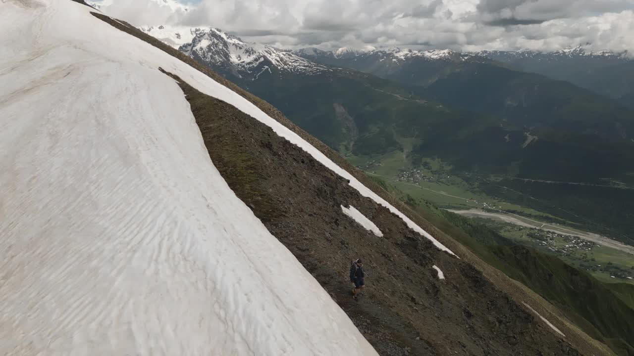 在积雪覆盖的高山中，背着背包的徒步旅行者的航拍照片。多云的天空下，一个人走在陡峭的岩石小道上。徒步旅行者探索自然，享受冒险。美丽的山景。视频素材