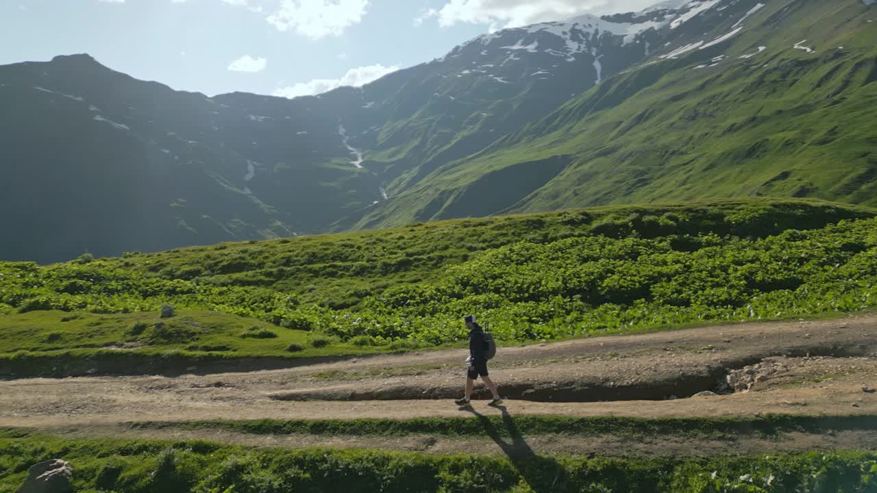 背包客在高山草地徒步旅行的鸟瞰图。人沿着土路走，四周是青山、山峰、碧蓝的天空。户外探险、旅游、自然探索。无人机拍摄的视频素材