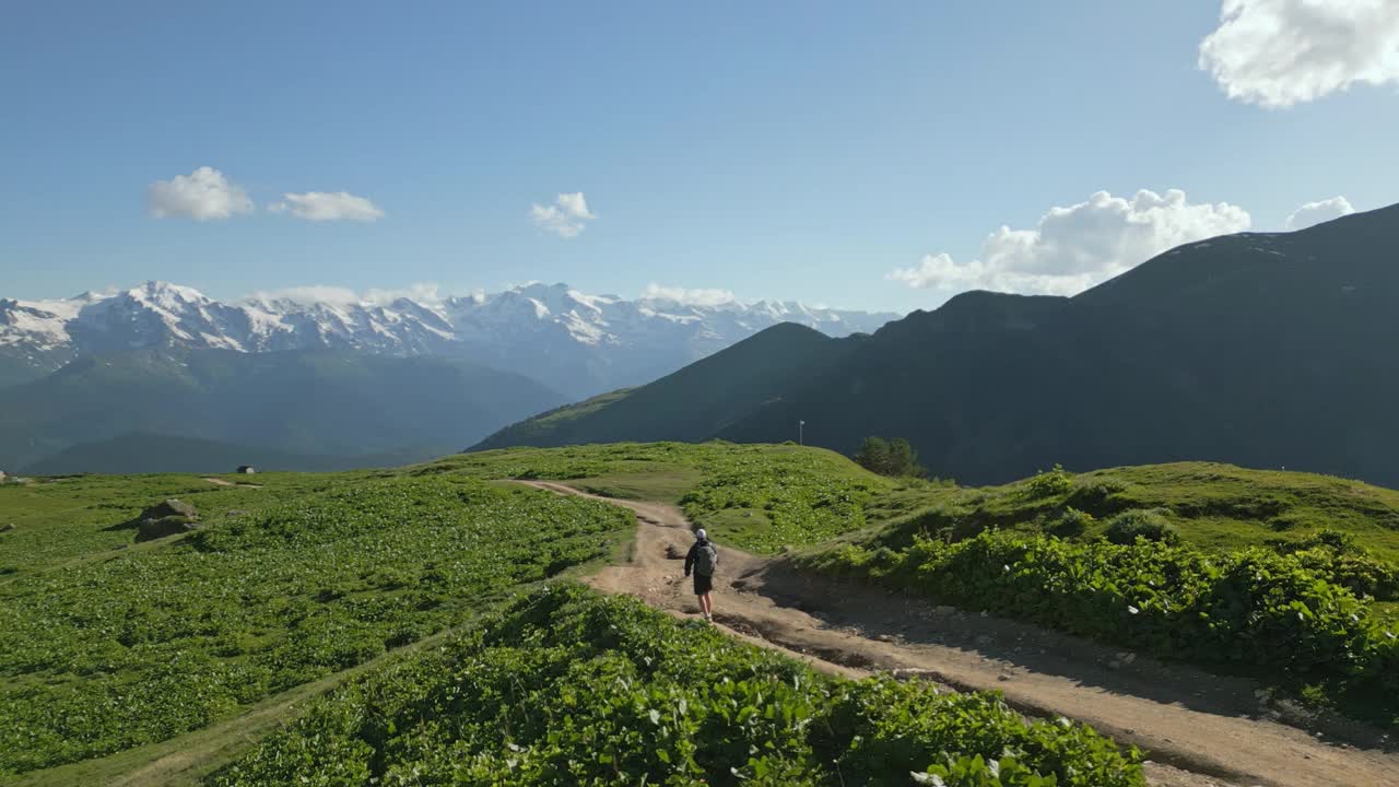 鸟瞰独自背包客在高山草地上行走。人们沿着崎岖的小路徒步旅行，周围是绿色的山丘，远处的雪峰。冒险、户外、探险主题。视频素材