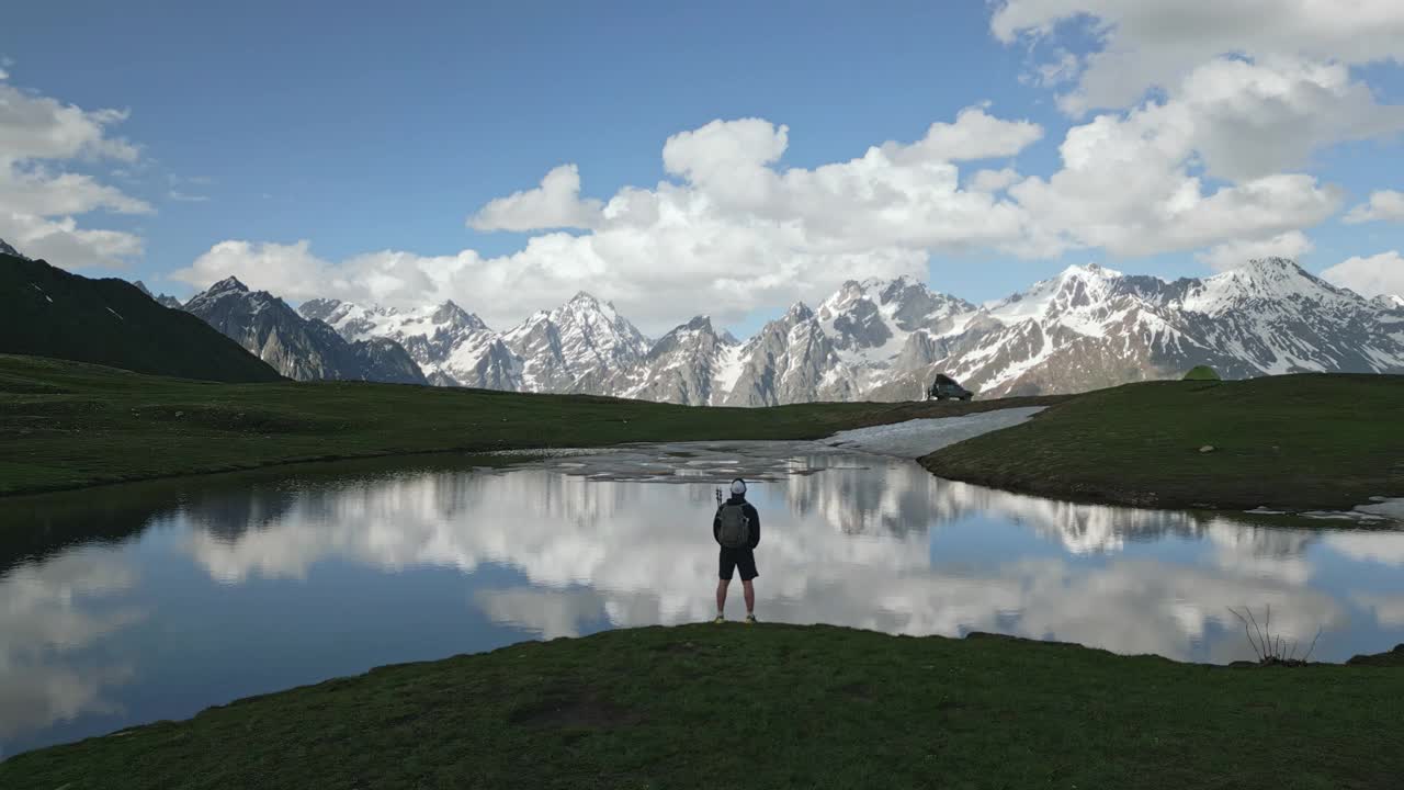 在斯瓦涅季山脉Koruldi湖附近，背着背包的徒步旅行者鸟瞰图。在林木线上，雪峰和云彩映照在湖面上。独自旅行者探索遥远的高山风光。无人机拍摄的视频素材