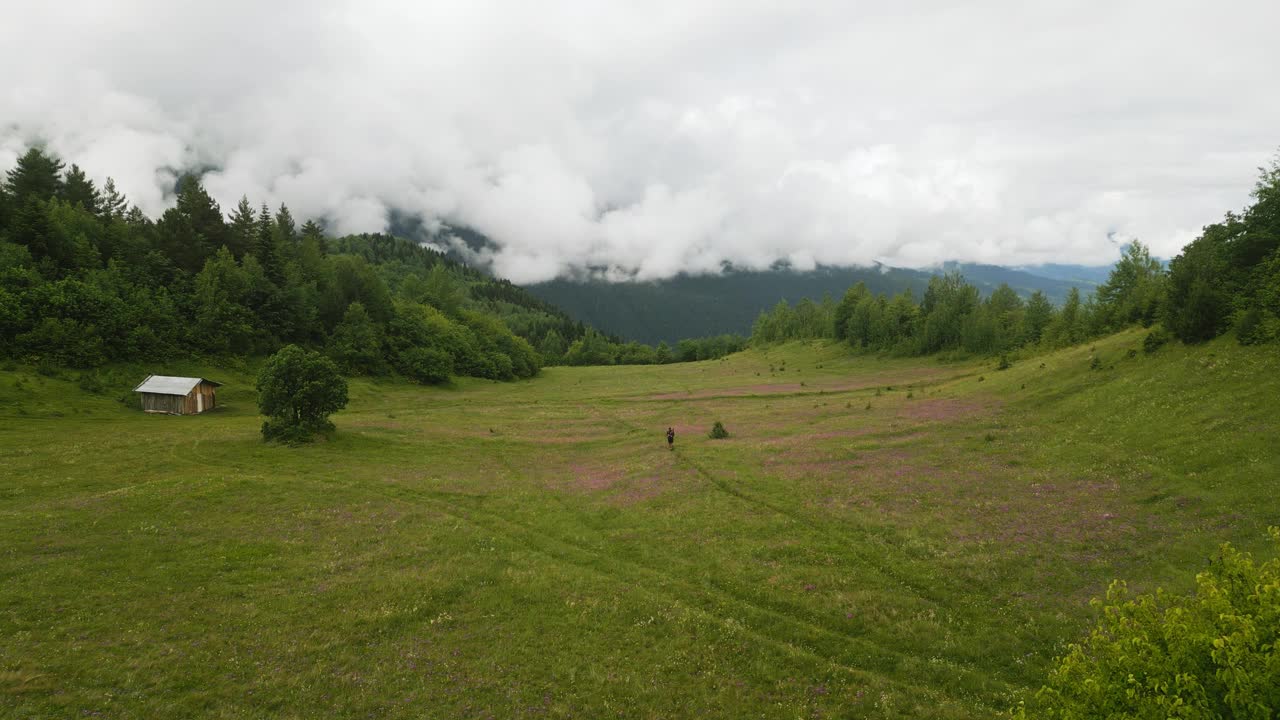 鸟瞰图独自背包客徒步旅行在高山草甸。人带着小木屋穿过绿野，靠近森林、山峦、云彩。徒步旅行、自然探险、探险、户外活动。视频素材