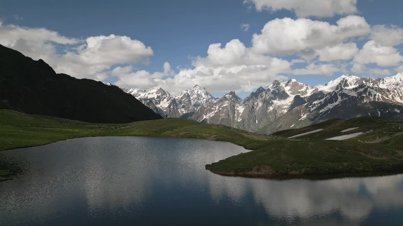 Koruldi山湖泊和Svaneti山峰的鸟瞰图。一个徒步旅行者走过。雪片和绿色的草地。风景优美的自然风光。夏季徒步旅行，户外探险无人机拍摄视频素材