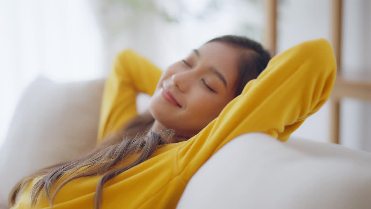 放松的年轻亚洲女人在舒适的沙发上享受休息，平静迷人的女孩在沙发上放松，闭上眼睛呼吸新鲜空气，在家冥想视频下载