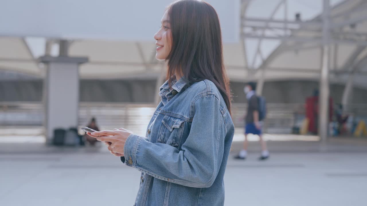 年轻的亚洲女子穿着牛仔裤，夹克，衣服，在晴朗的日子里走在城市的街道上，用智能手机发短信视频素材