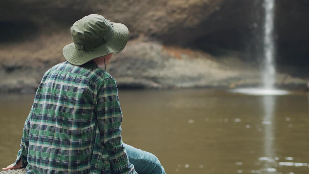 一个男孩坐在瀑布旁的岩石上。那个人戴着一顶绿色的帽子，穿着格子衬衫视频下载