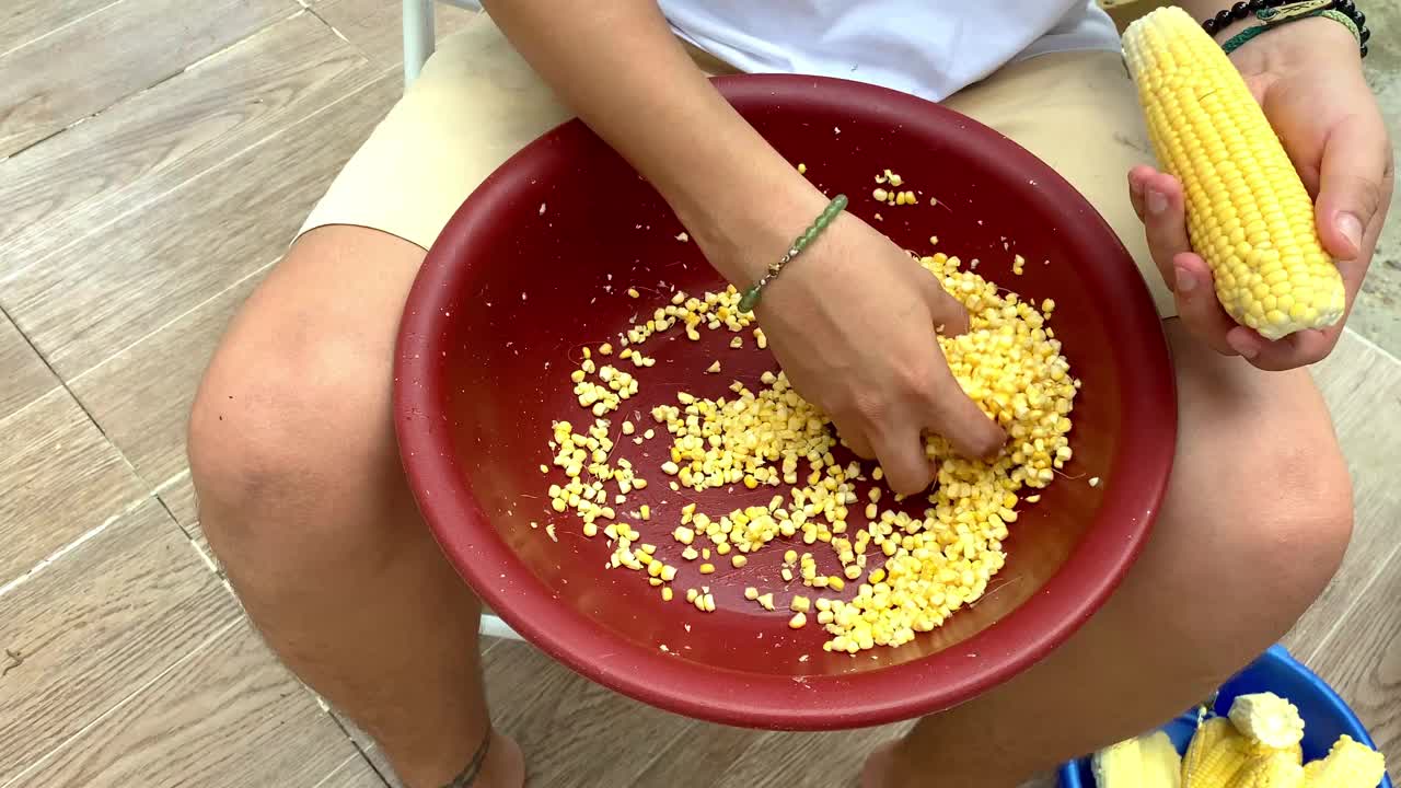 在户外，用手把新鲜玉米剥到一个红碗里的特写镜头。视频下载