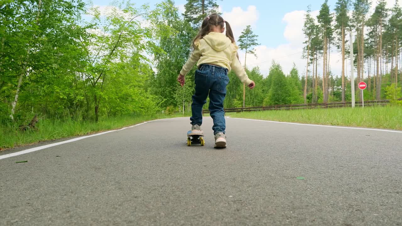 小女孩在公园里的小路上慢慢地滑滑板，这是一个阳光明媚的夏日，慢动作。视频素材