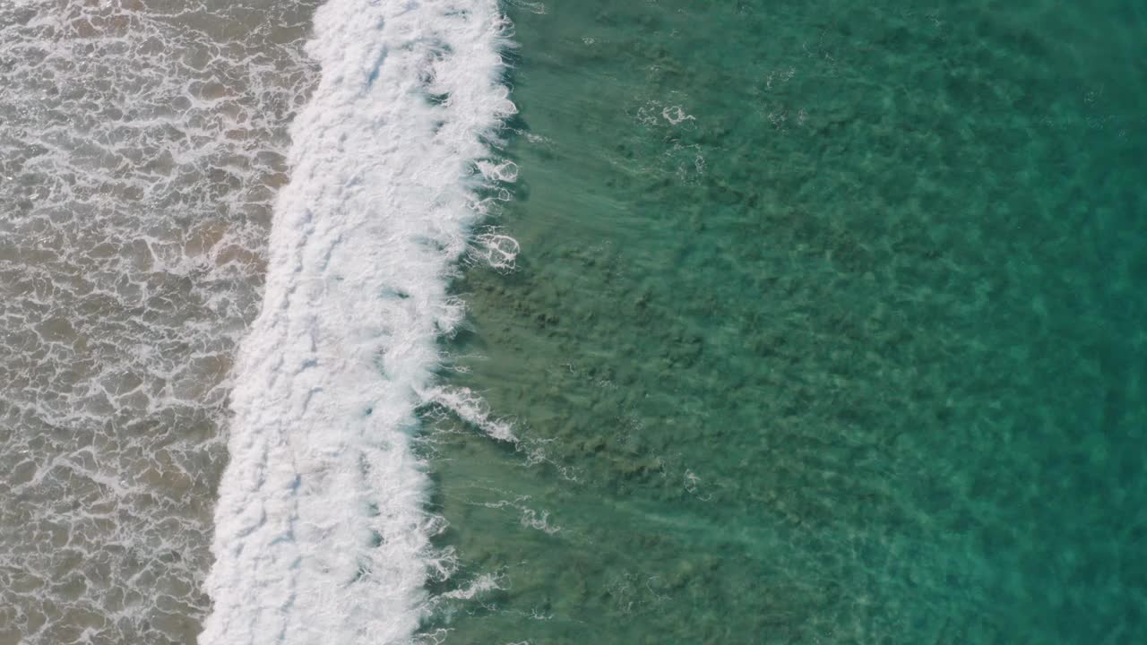 鸟瞰美丽的绿松石水波沿着田园诗般的清澈，浅，沙质海岸线破碎，创造抽象的移动海泡沫纹理视频下载