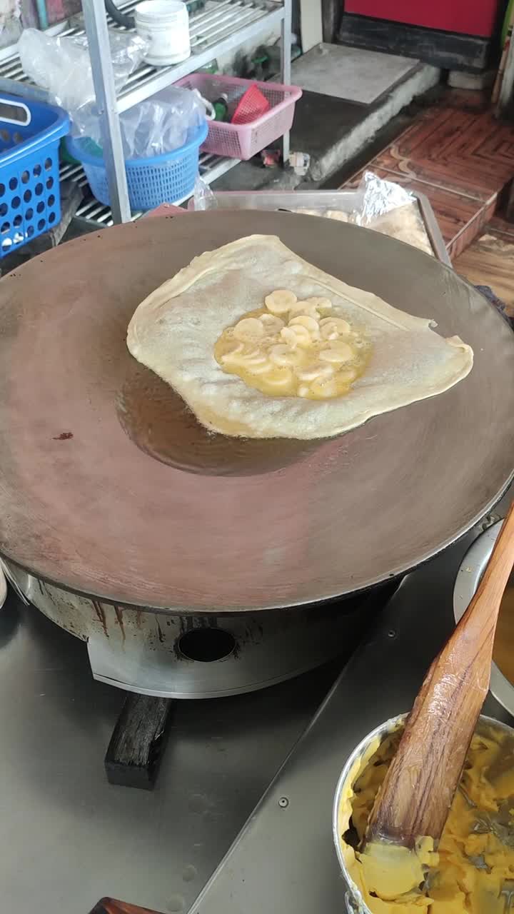 在露天市场，一个街头小贩在热煎锅上准备香蕉薄饼，展示了传统的烹饪方法。视频下载