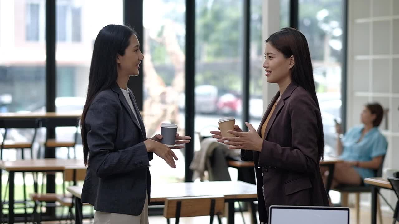 两个穿西装的女人在咖啡馆里聊天。其中一个拿着杯子。另一个女人拿着一个钱包视频下载