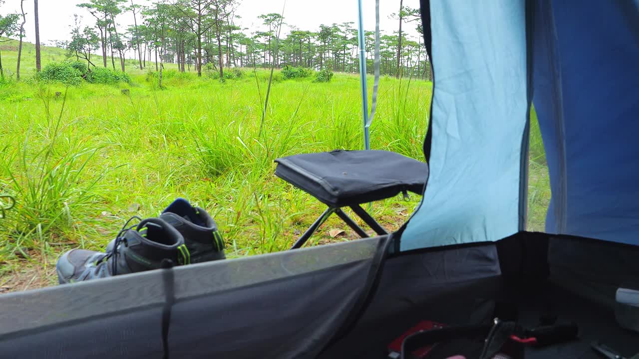 露营的早晨，从帐篷里，可以看到远处一片宁静的草地。登山靴，野营椅，以及用于探险和户外生活的便携式炉子。视频下载