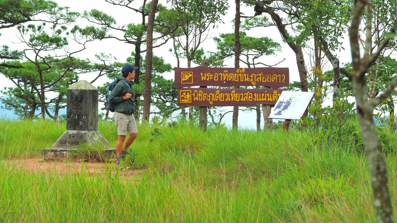 一名亚洲男子背着双肩包在泰国Phu Soi岛签名。四周绿草如茵，绿树成荫。指示牌指示前往“富索岛日出”及“双板界碑”的方向。视频素材