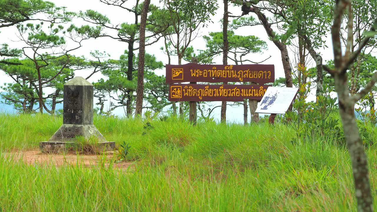 一名亚洲男子背着双肩包在泰国Phu Soi岛签名。四周绿草如茵，绿树成荫。指示牌指示前往“富索岛日出”及“双板界碑”的方向。视频素材