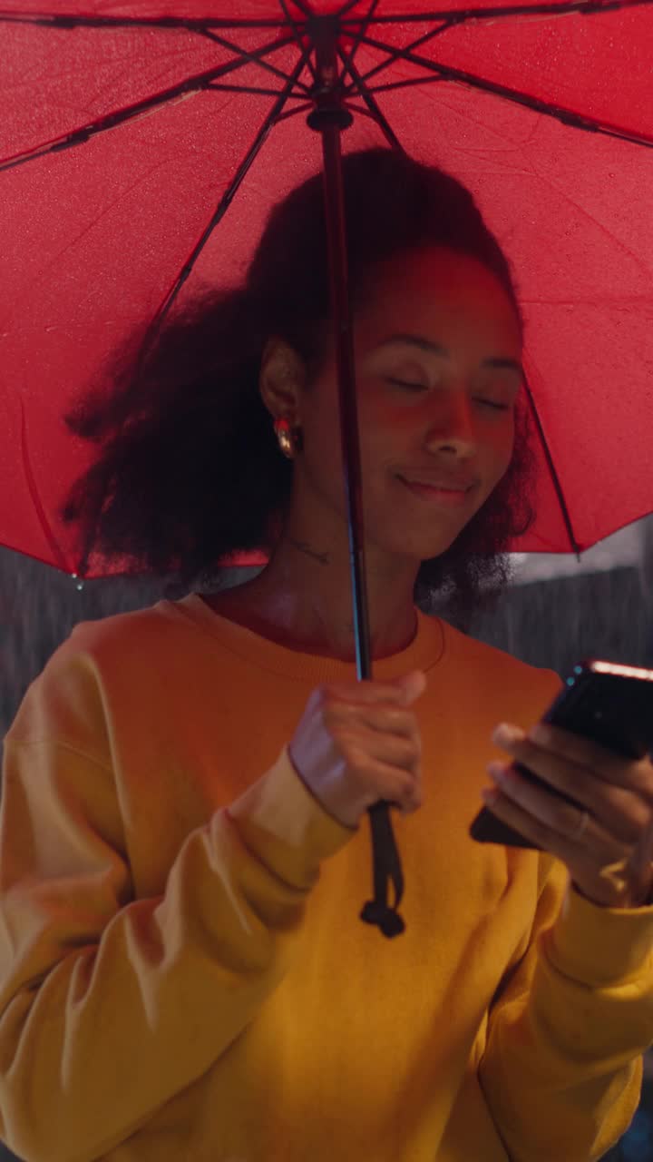 垂直屏幕:一个下雨天，年轻的黑人女性在一把红色雨伞下微笑着使用智能手机。女孩浏览社交媒体应用，发短信，站在一个城市的夜晚视频素材