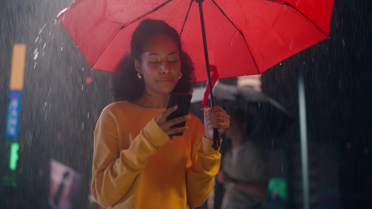 一个非洲女孩站在大雨滂沱的夜里，一边浏览社交媒体应用，一边给朋友发短信。穿着黄色衣服的年轻快乐的黑人女性微笑着使用她的智能手机视频素材