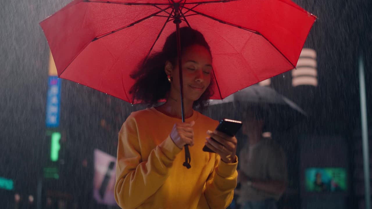 一个下雨天，穿着黄色毛衣的年轻快乐的黑人女性在一把红色雨伞下微笑着使用智能手机。女孩浏览社交媒体应用，发短信，站在一个城市的夜晚视频素材