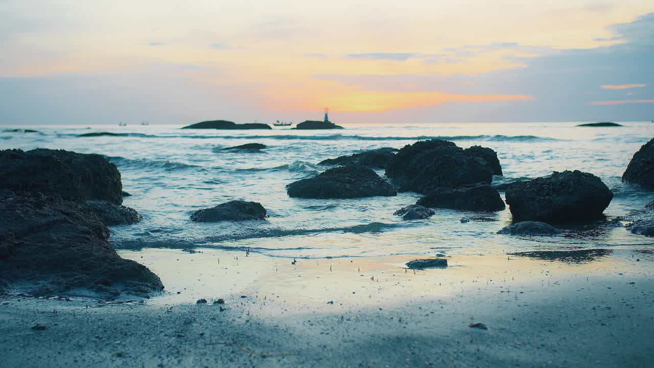 日落时宁静的海岸景色，有岩石海岸线，平静的海浪，远处的灯塔在五彩缤纷的天空下。视频素材