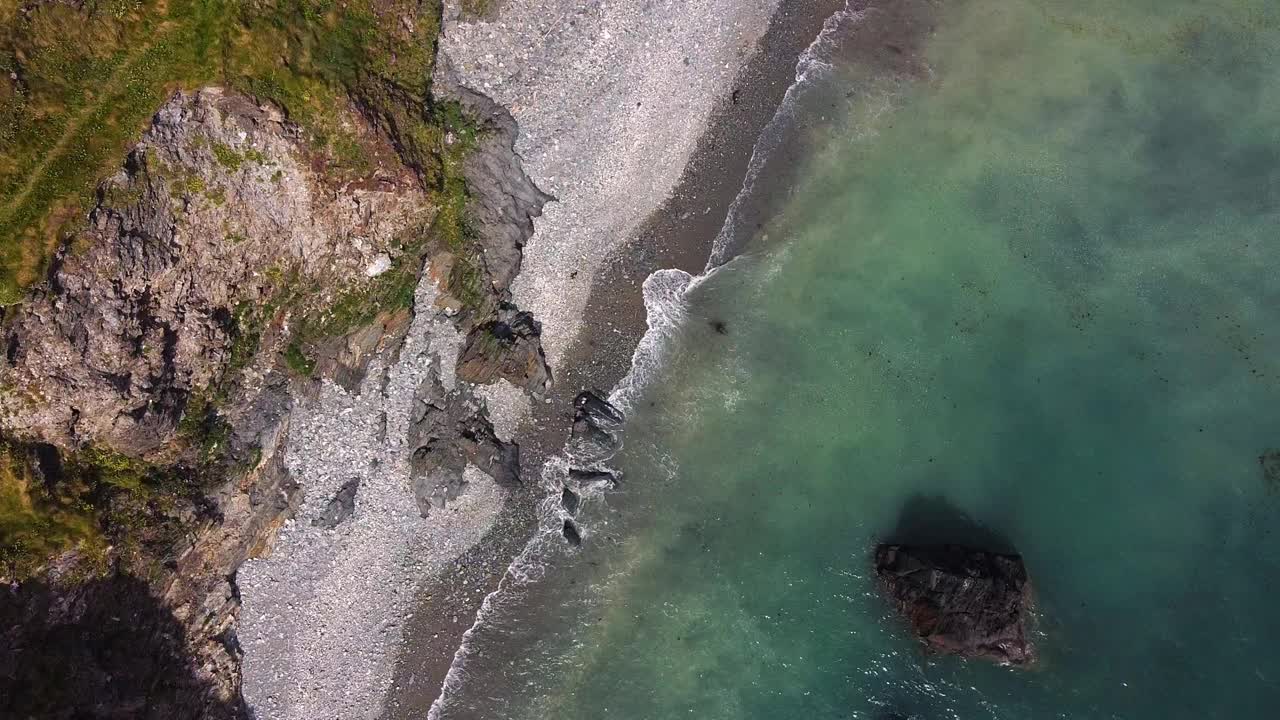 岩石海岸线的鸟瞰图。海水清澈湛蓝，海滩上布满了小石头。一大块岩石伸出水面。视频素材