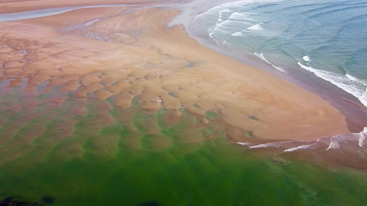 低潮时，从空中拍摄的波浪沿着沙滩海岸线破碎。海浪在海滩上形成了复杂的图案，展示了海岸线上水和陆地的美丽混合。视频素材