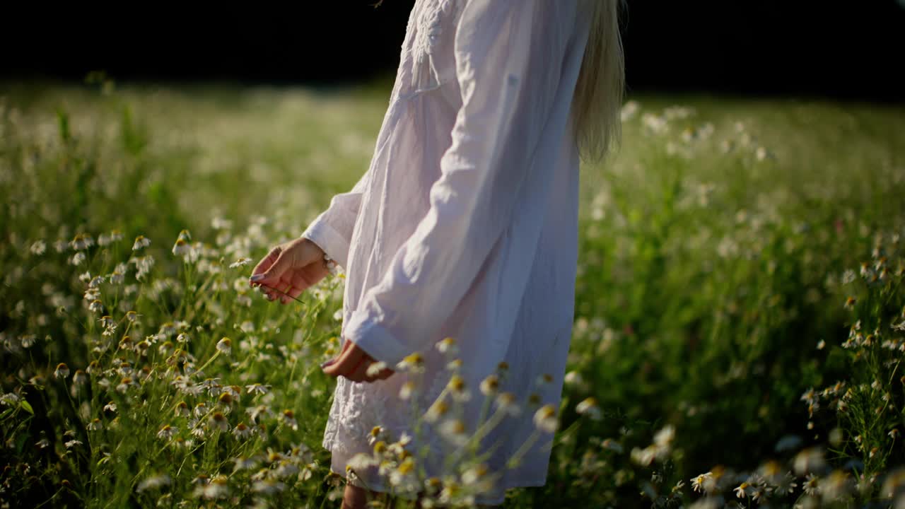 在阳光明媚的草地上，身穿白色连衣裙的女子欣赏鲜花视频素材