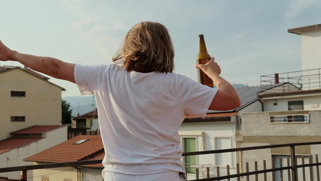 快乐的女人拿着一瓶啤酒在屋顶上跳舞在日落时分喝啤酒视频素材